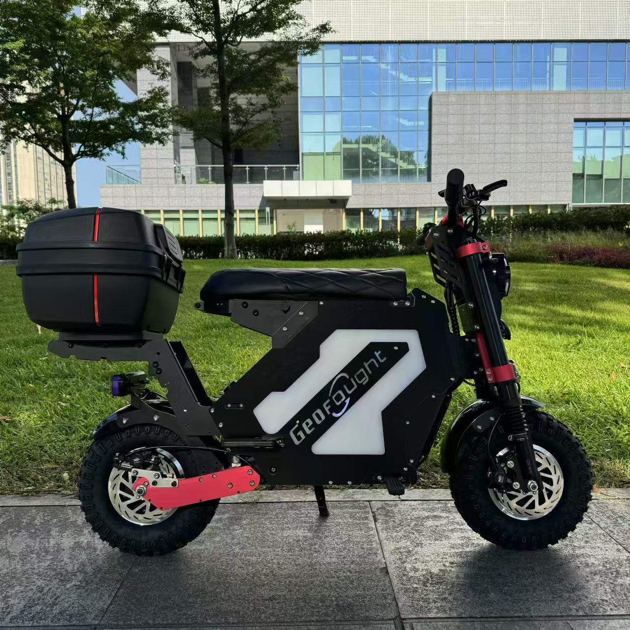A front view of a mini electric motorcycle designed for off-road use, equipped with a rear trunk, showcased in an urban park setting.