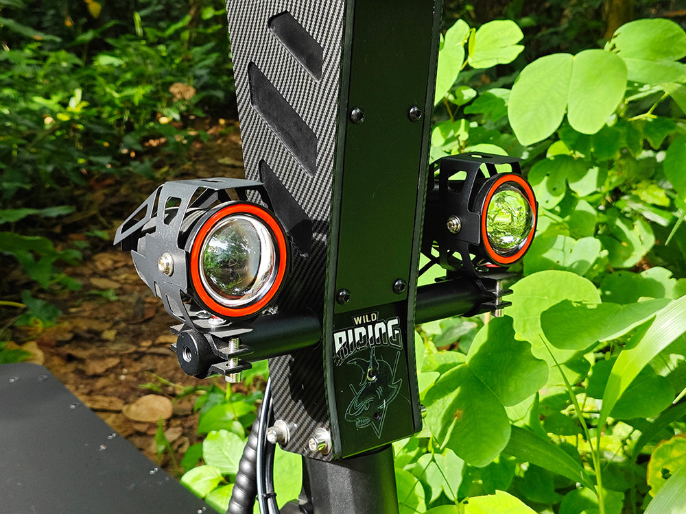 Close-up of dual powerful LED headlights mounted on an off-road electric scooter, surrounded by lush greenery. The rugged design highlights the scooter’s capability for off-road adventures.