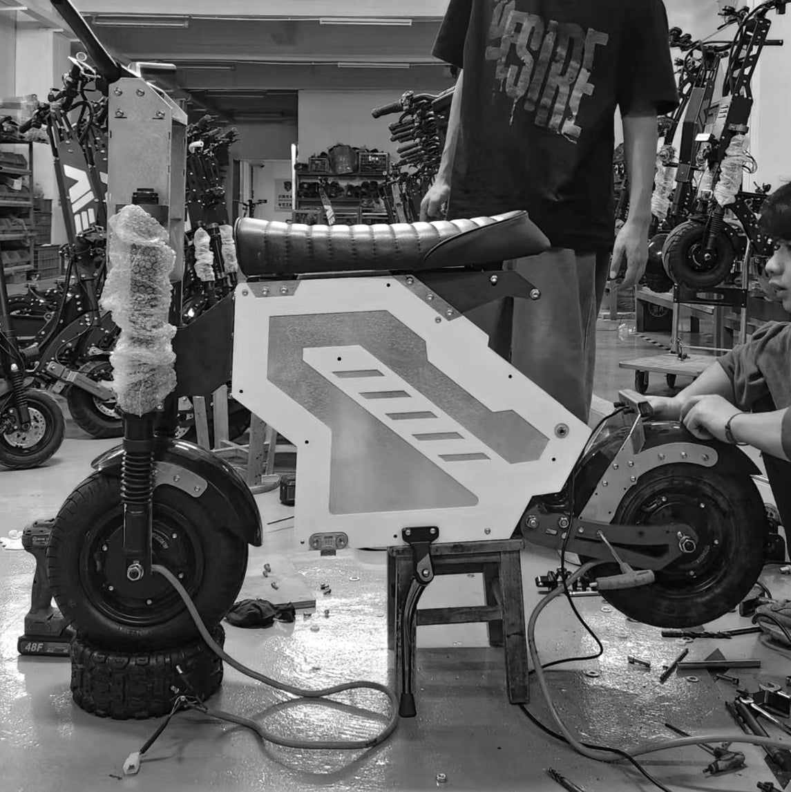 Workers assembling an electric motorbike in a factory setting. The bike is elevated on a stand, with various components being installed, highlighting the production process.