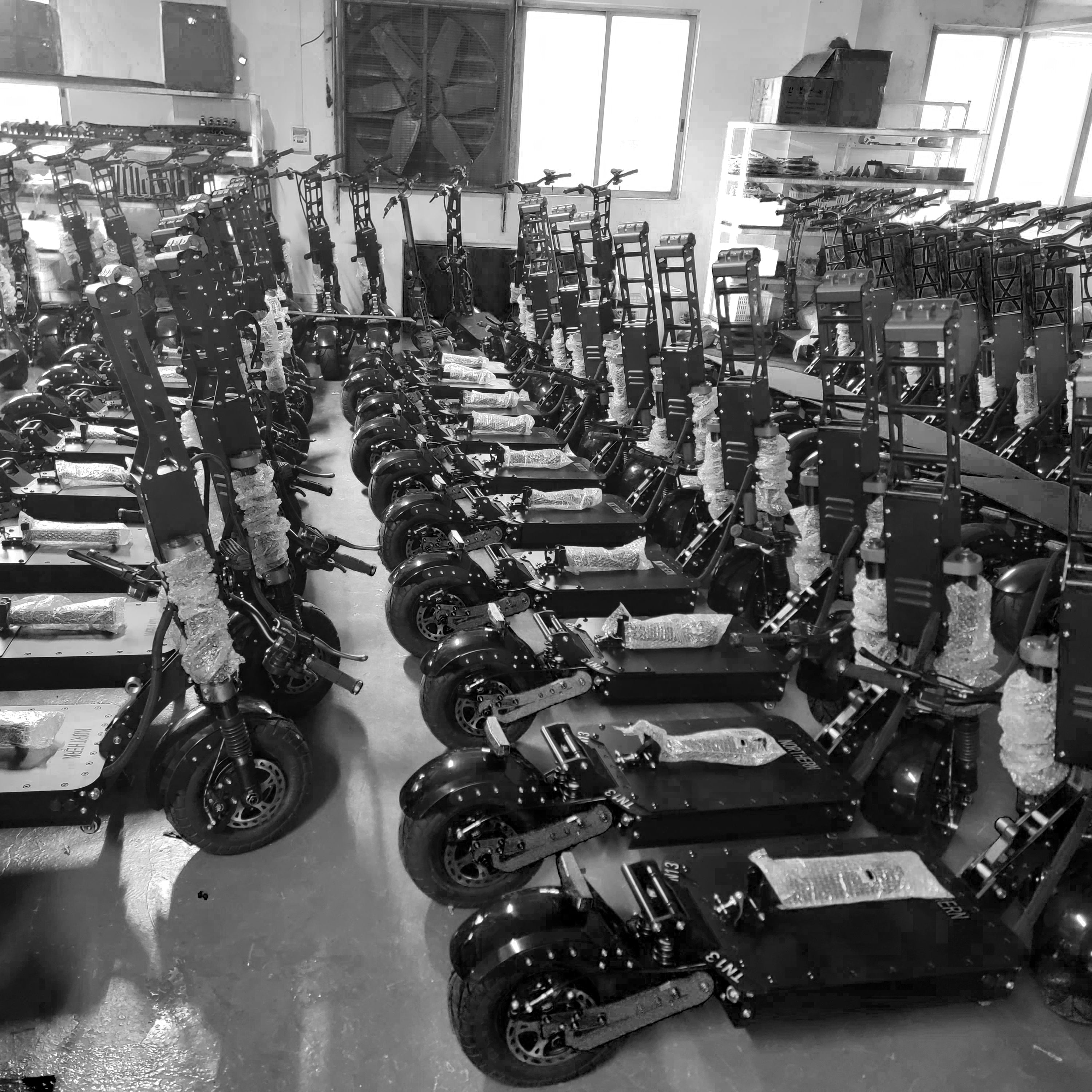 A large number of electric scooters neatly lined up in a factory storage room, ready for packaging and distribution. The scooters are partially assembled and protected with packaging material.