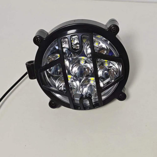 High-intensity LED headlight for electric motorcycles. The headlight is wrapped in a durable black protective grille to ensure safety while enhancing the rugged aesthetic of the vehicle. It features multiple LED bulbs configured in a circular pattern to provide a wide and bright beam, essential for enhanced nighttime visibility and road safety.