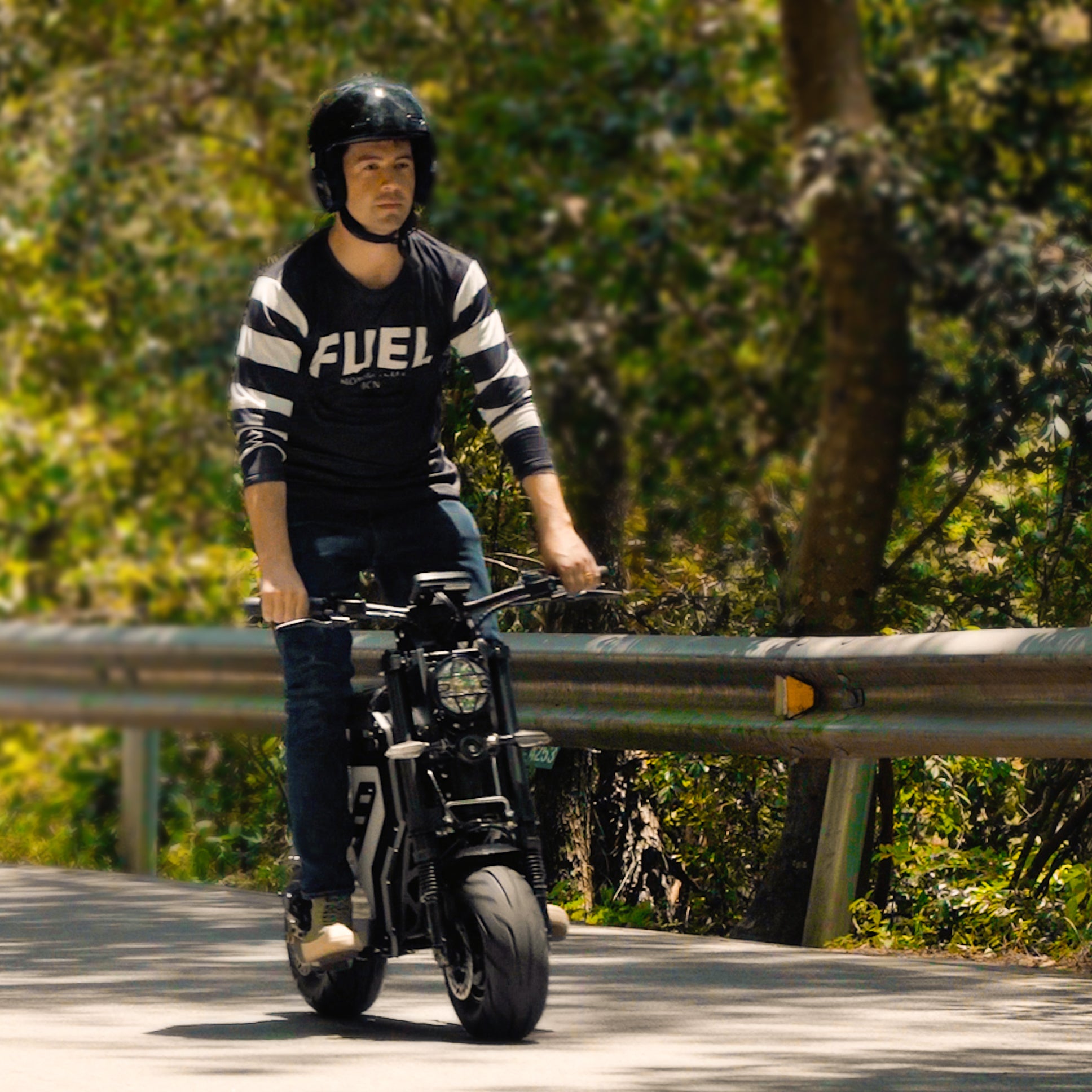 A man riding an electric motorbike on a winding forest road. He is wearing a helmet and a "FUEL" sweatshirt, highlighting the bike's capability for smooth handling on scenic routes.